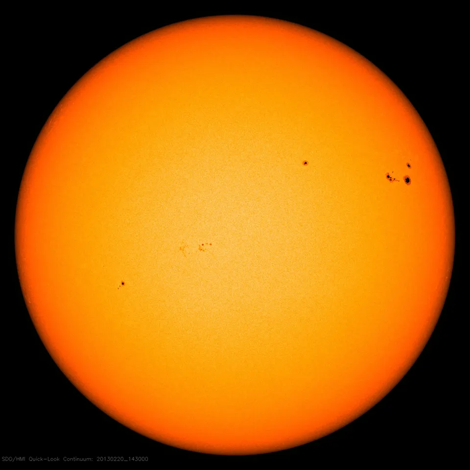 The Sun's full disc in visible light, bright at the centre and darker toward the edge, with a few small dark sunspot groups