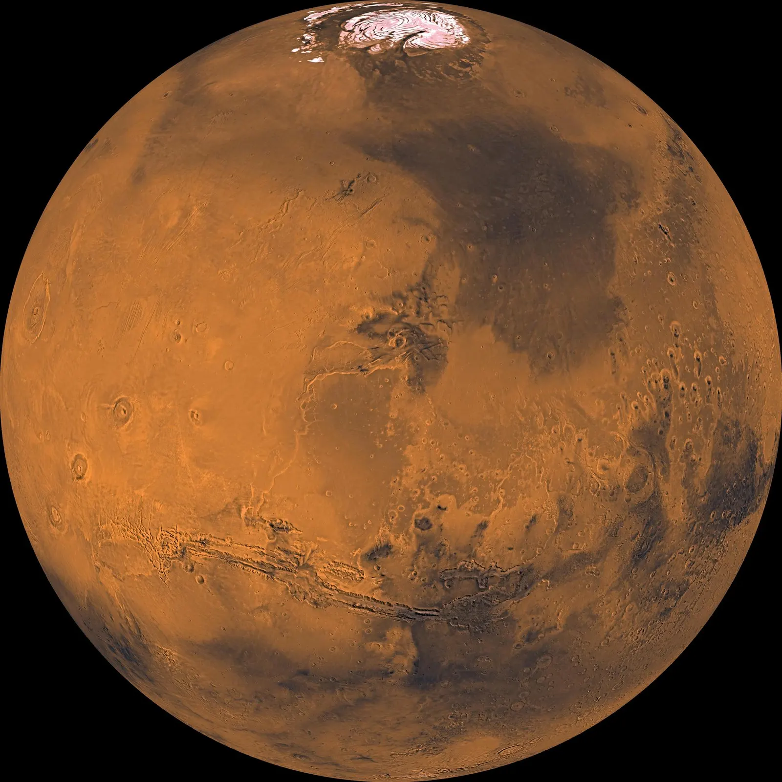 Mars as a red-orange globe crossed by the long dark canyon system of Valles Marineris, with volcanoes to the left.